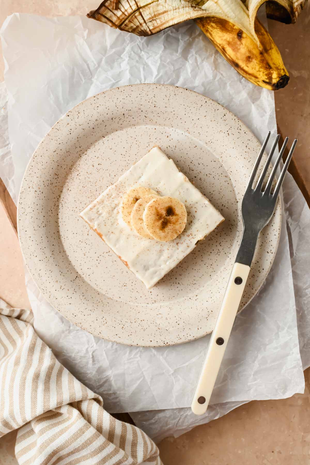 slice of banana sheet bake topped with cream cheese frosting and sliced bananas on speckled plate.