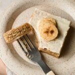 slice of banana sheet bake topped with cream cheese frosting and sliced bananas on speckled plate.