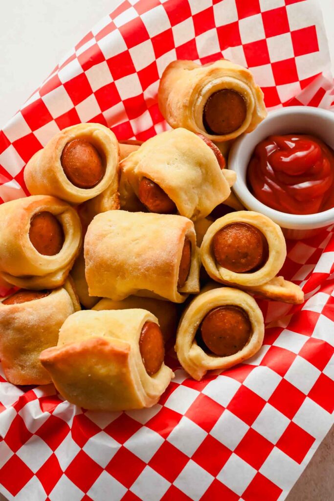 pigs in a blanket in basket lined with red and white checkered paper with small bowl of ketchup on side.
