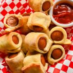 pigs in a blanket in basket lined with red and white checkered paper with small bowl of ketchup on side.