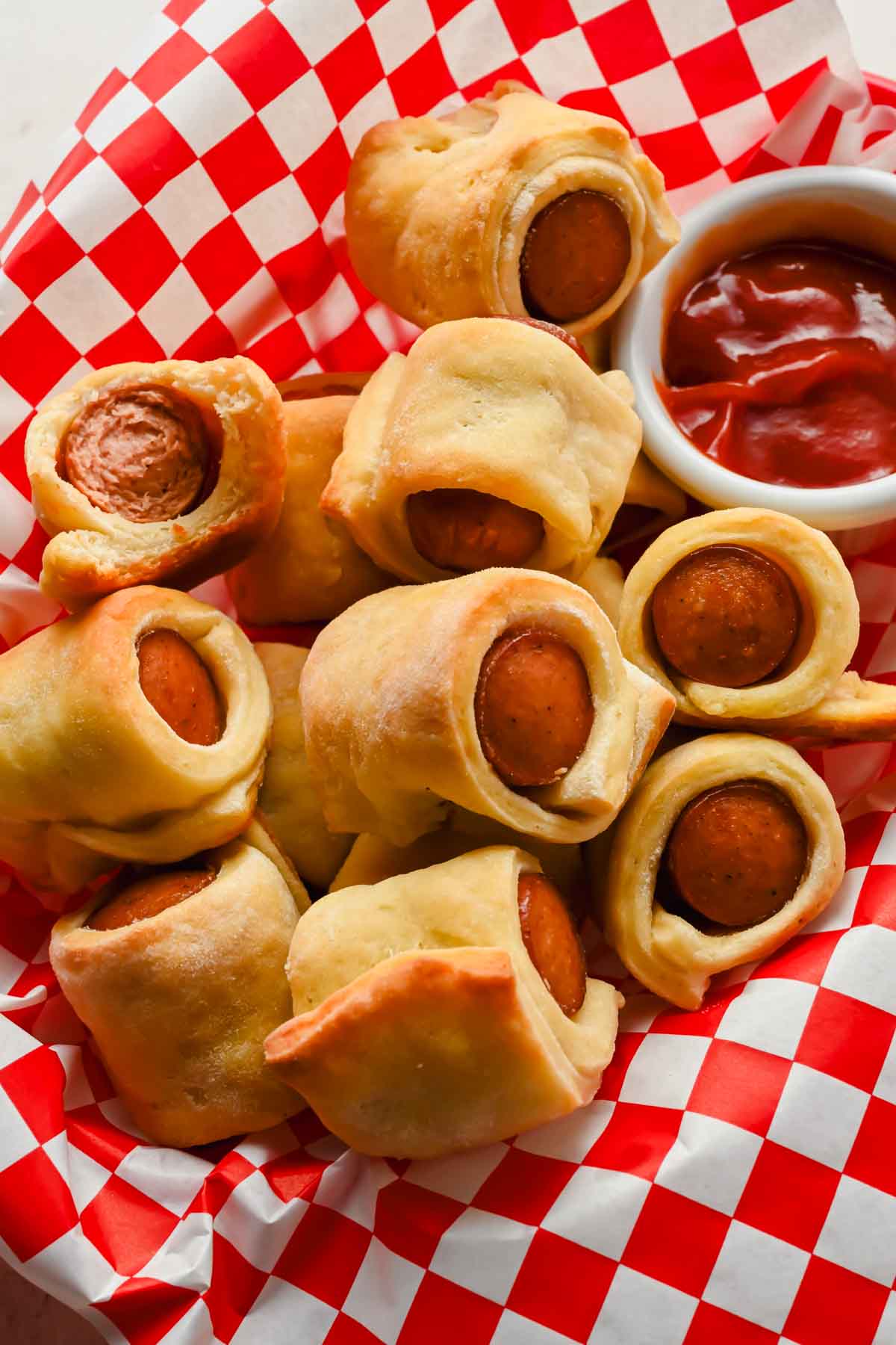 pigs in a blanket in basket lined with red and white checkered paper with small bowl of ketchup on side.