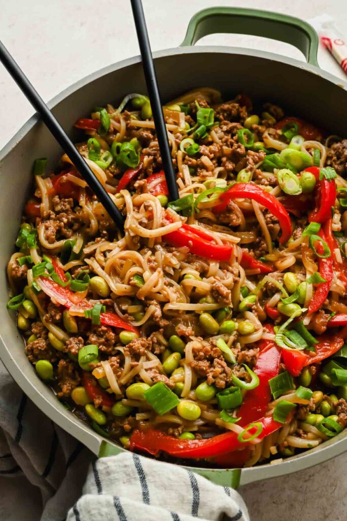 korean beef stir fry noodles in green ceramic pan topped with diced green onions with chopsticks in pan.