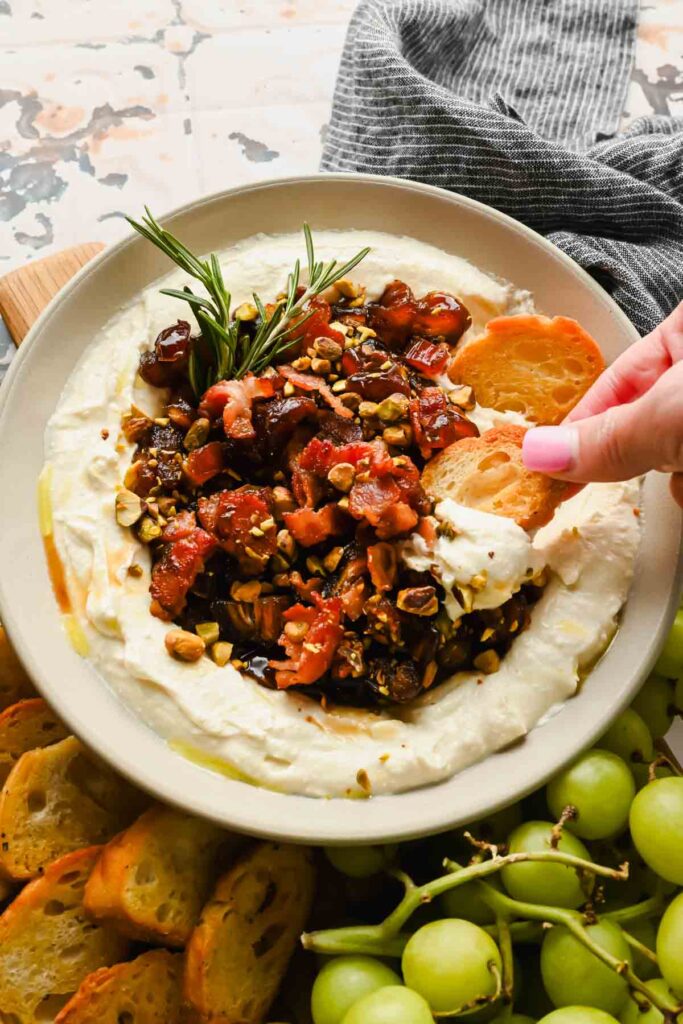 serving bowl with whipped feat cheese topped with a date, bacon, pistachio mixture garnished with rosemary.