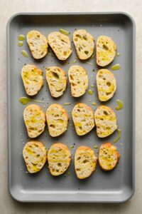 crostinis on gray sheet pan drizzled with olive oil, salt and pepper.