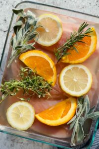 4 turkey tenderloins lined up in glass baking dish drowned in salt water brine topped with herbs, lemon slices and orange slices.