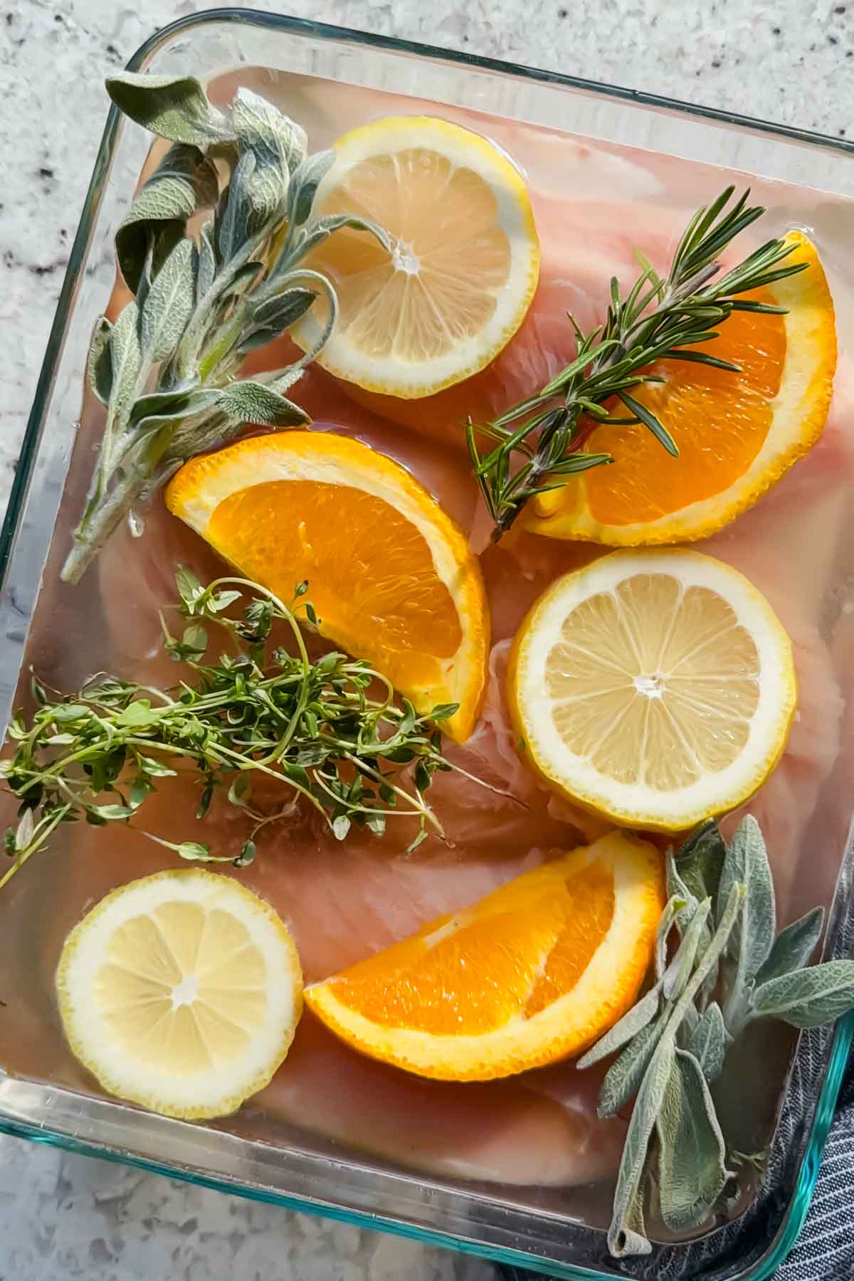 4 turkey tenderloins lined up in glass baking dish drowned in salt water brine topped with herbs, lemon slices and orange slices.