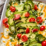 white pan of breakfast enchiladas topped with tomatoes, avocado, cheese and a creamy green sauce.