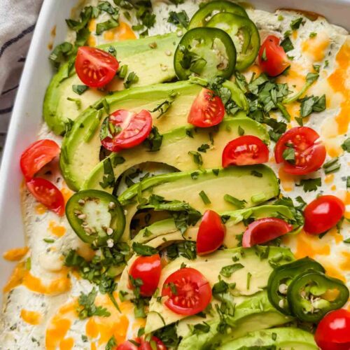 white pan of breakfast enchiladas topped with tomatoes, avocado, cheese and a creamy green sauce.