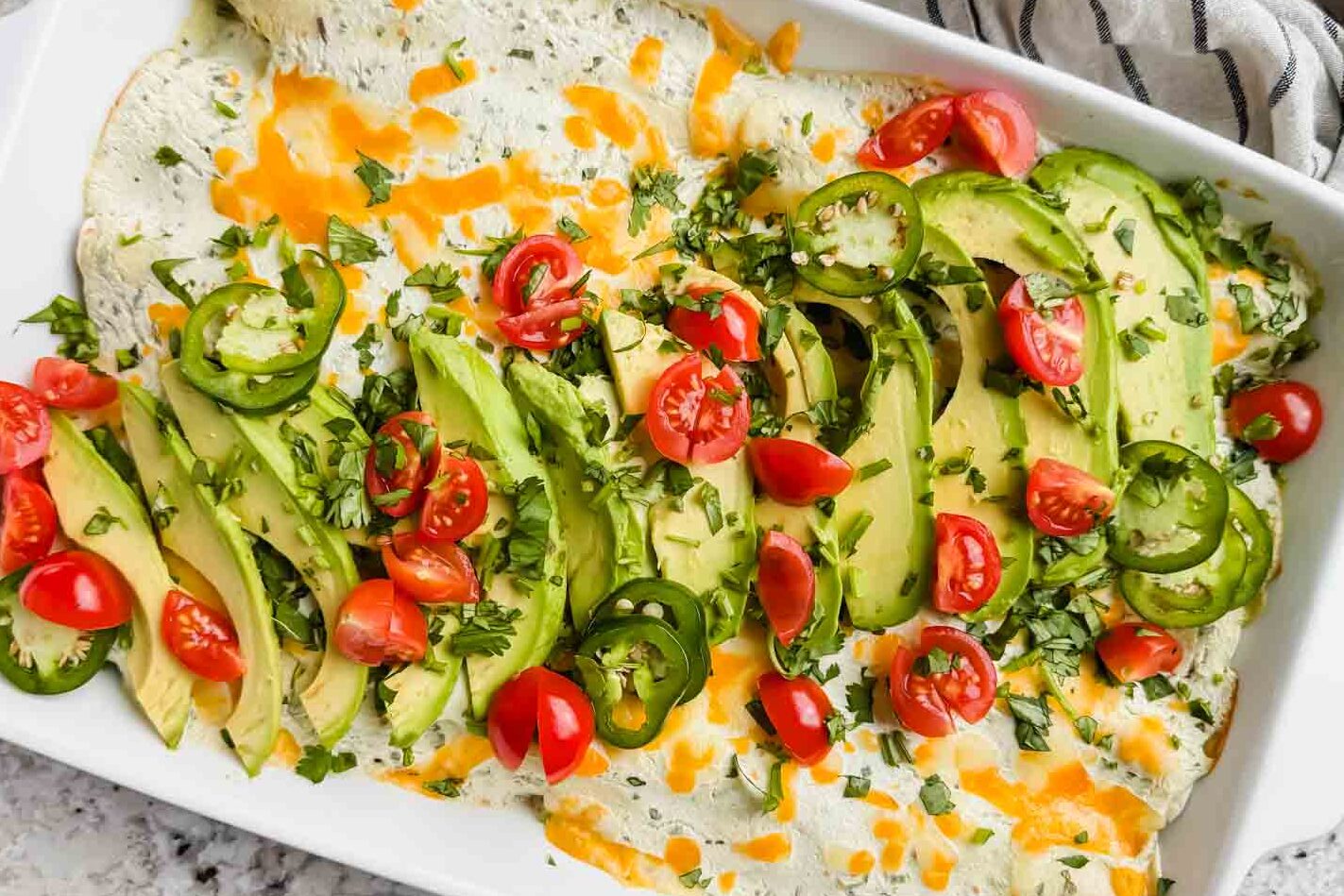 white rectangular dish filled with breakfast enchiladas topped with avocado, tomatoes, cheese and a green sauce.