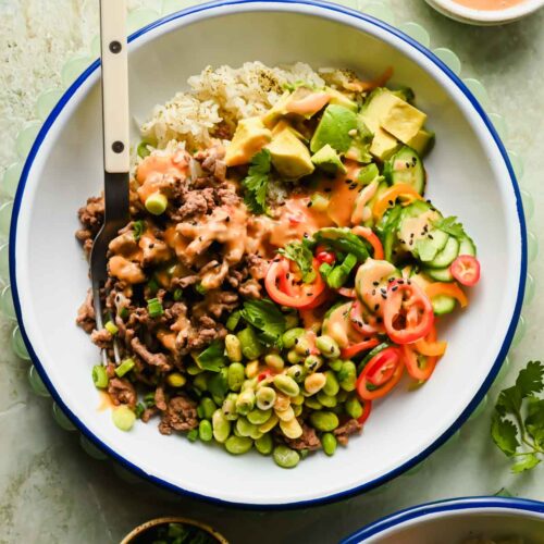 ground beef bang bang bowl with sliced avocado, edamame and bell peppers drizzled in sauce.
