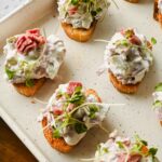 crostinis topped with corned beef dip and sprouts on white baking sheet.