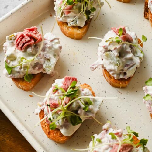 crostinis topped with corned beef dip and sprouts on white baking sheet.