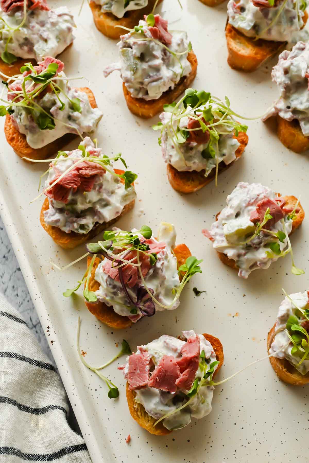 crostinis topped with corned beef dip and sprouts on white baking sheet.
