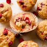 cranberry apple muffin with muffin liner peeling off surrounded by additional muffins on parchment.
