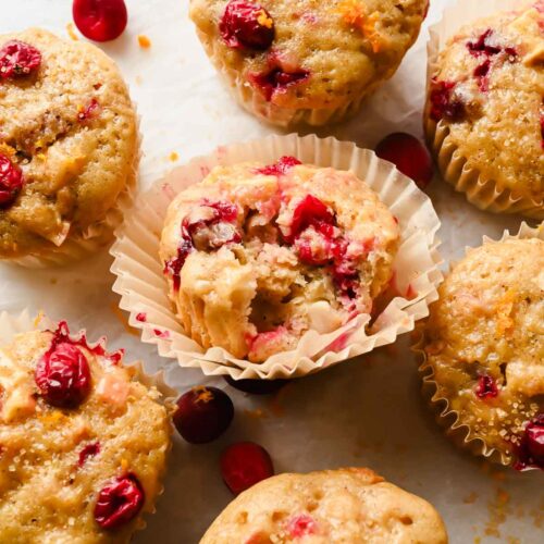 cranberry apple muffin with muffin liner peeling off surrounded by additional muffins on parchment.