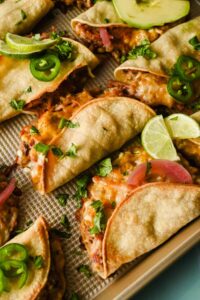 baked crispy pork tacos topped with herbs, lime wedges, pickled onions, and avocado.