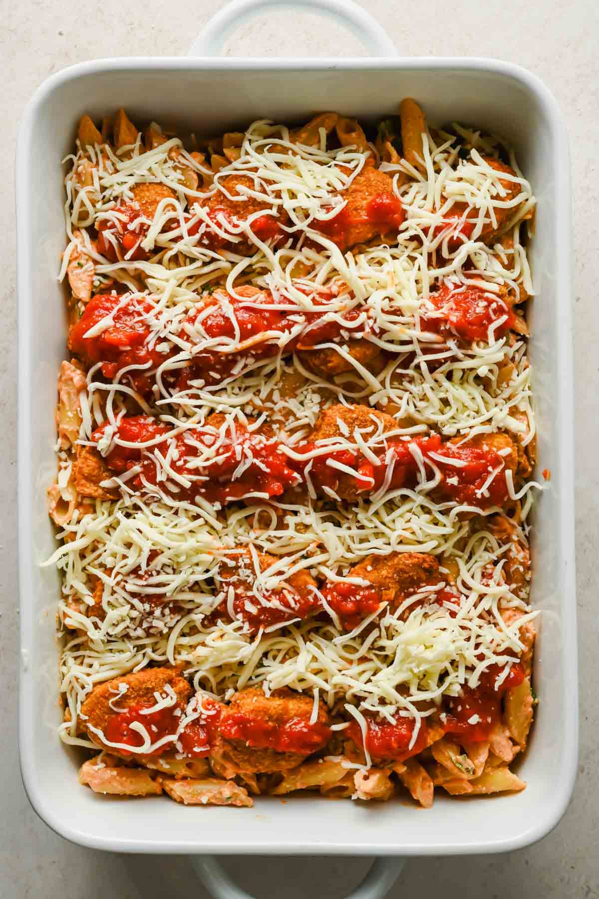 healthier chicken parm bake in rectangular baking dish topped with shredded mozzarella before baking.