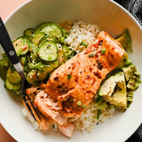 spicy salmon on top of bed of rice plated with avocado and side of cucumber salad.