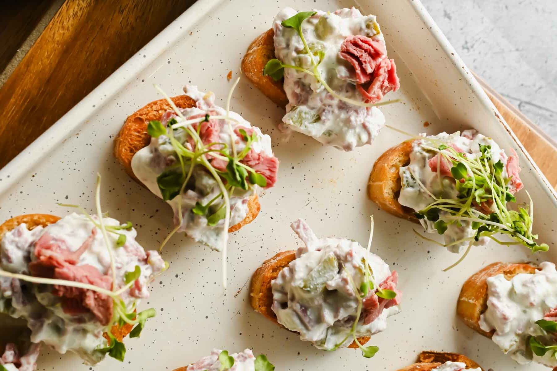 crostinis topped with corned beef dip and sprouts on white baking sheet.