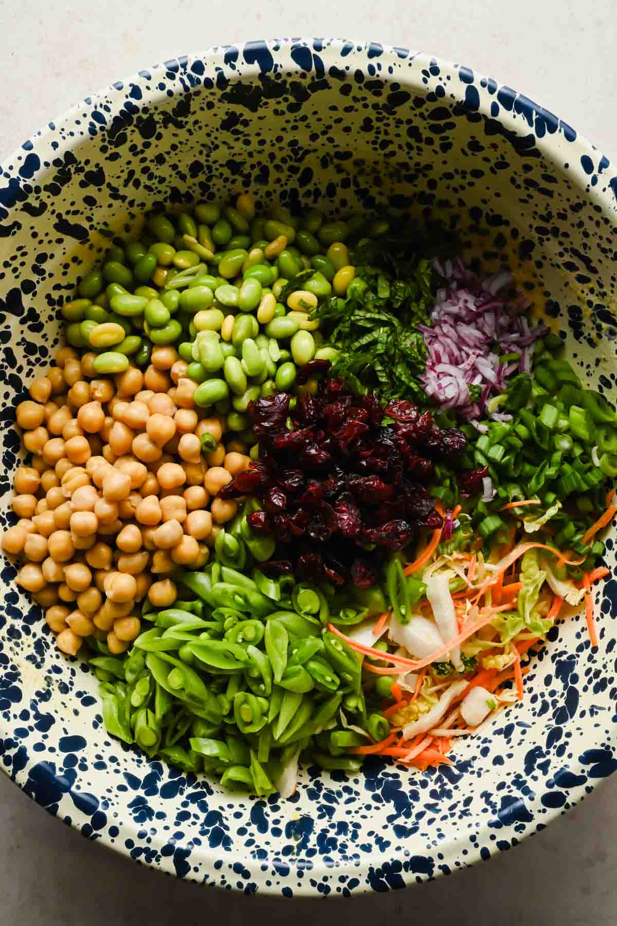 sliced snap peas, edamame, cabbage and carrot slaw, chickpeas and dried cranberries in large bowl.