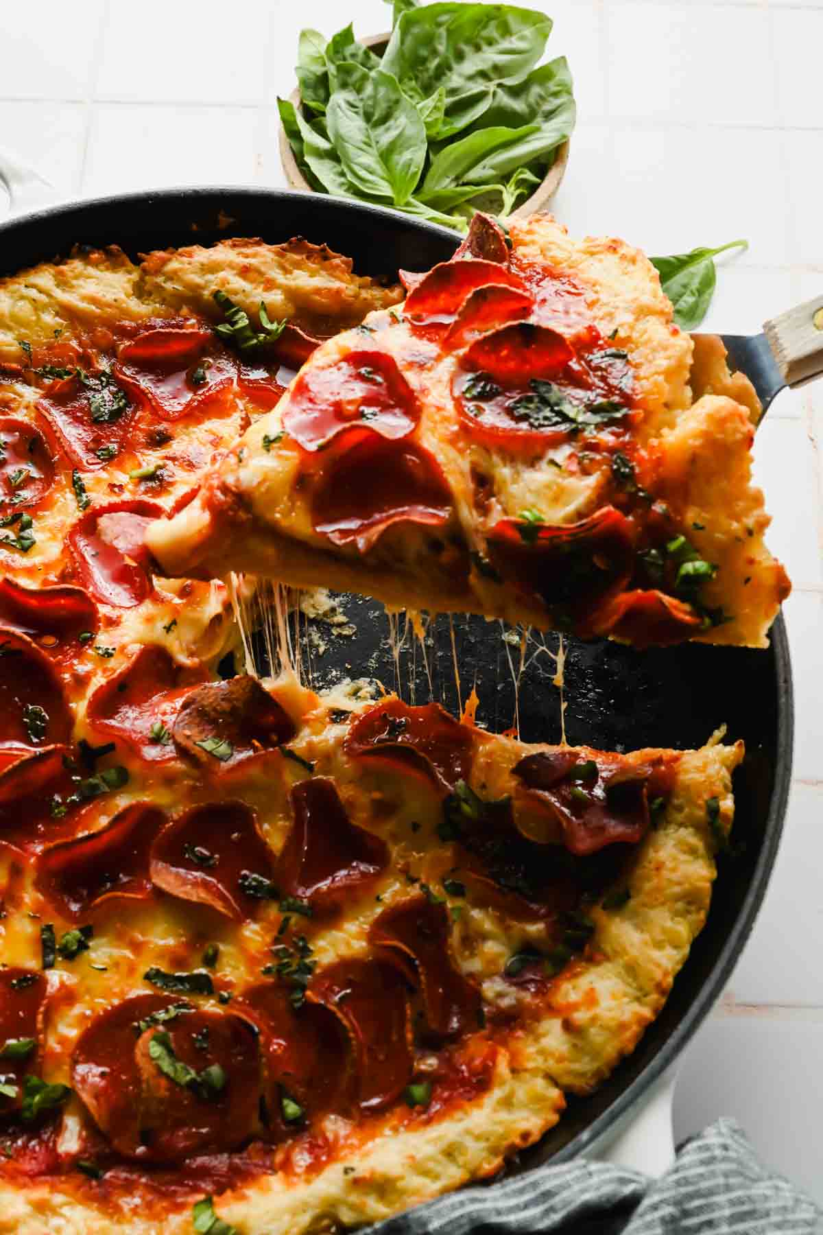 spatula taking slice out of skillet pizza topped with pepperoni, melty cheese and fresh herbs.