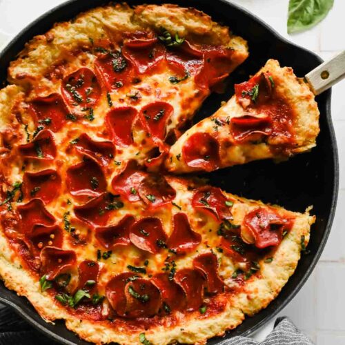 spatula taking slice out of skillet pizza topped with pepperoni, melty cheese and fresh herbs.
