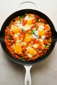 diced veggies and thick tomato sauce spread across cast iron skillet with 6 eggs cracked into pan and topped with fresh basil.