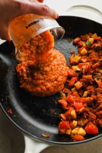tomato sauce being poured into cast iron skillet with cooked veggies.