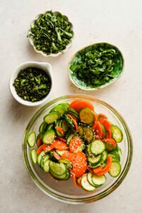 fresh herbs diced in small bowls beside larger bowl of carrot cucumber salad.