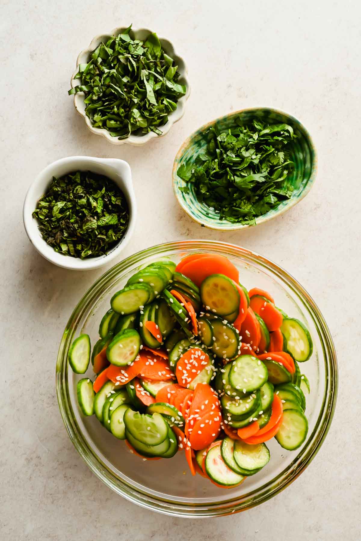 fresh herbs diced in small bowls beside larger bowl of carrot cucumber salad.