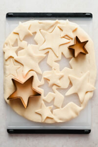 pie dough rolled out on cutting board cut into star shapes with cookie cutters.