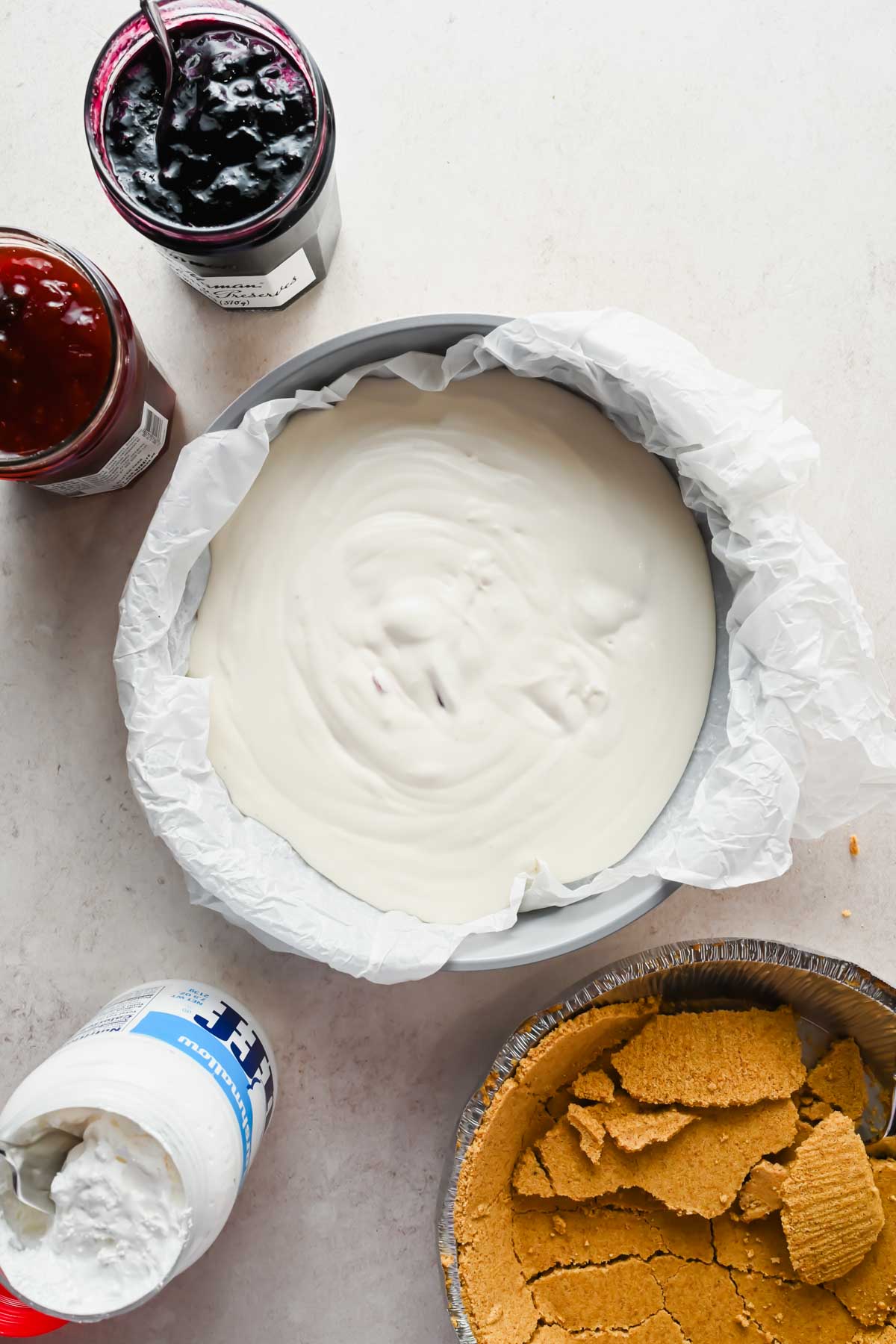 ice cream base in pie dish beside jar of berry preserves, marshmallow fluff and graham cracker crust.