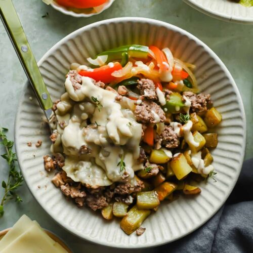 Philly cheesesteak bowl on white plate topped with cheese sauce and fresh herbs.