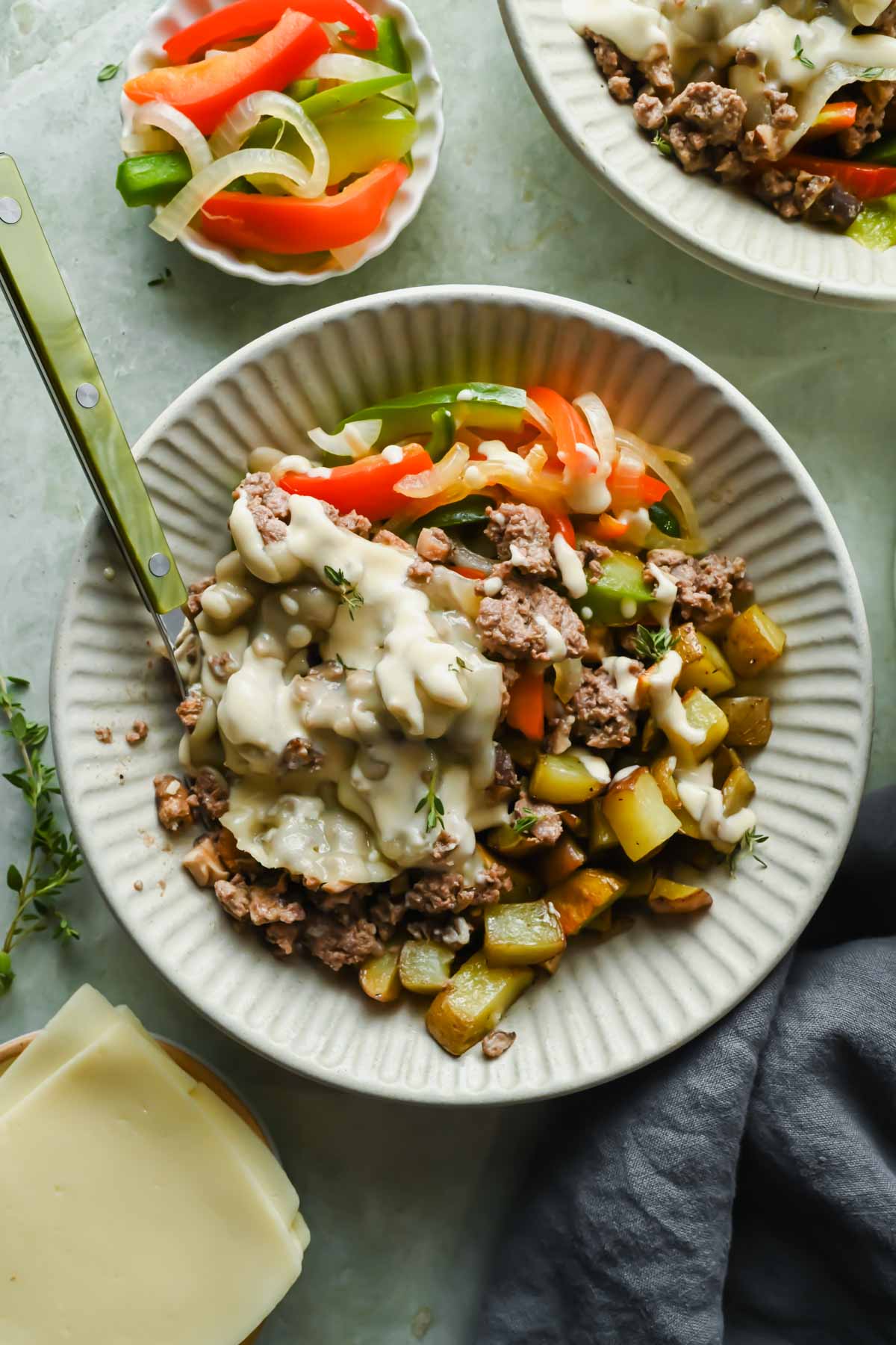 Philly cheesesteak bowl on white plate topped with cheese sauce and fresh herbs.