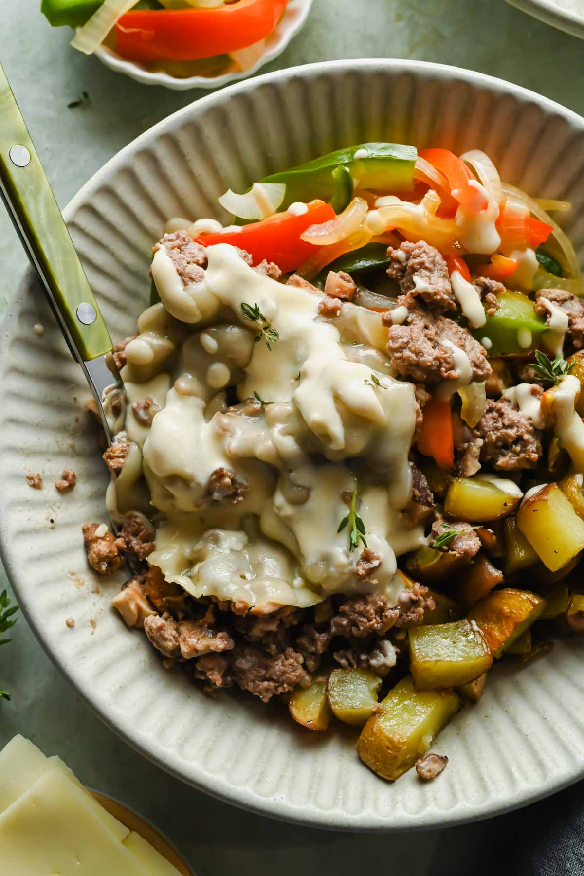 Philly cheesesteak bowl on white plate topped with cheese sauce and fresh herbs.
