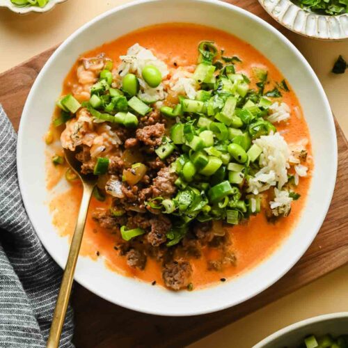 ground beef coconut rice bowl topped with edamame and fresh herbs in white serving dish with gold spoon.