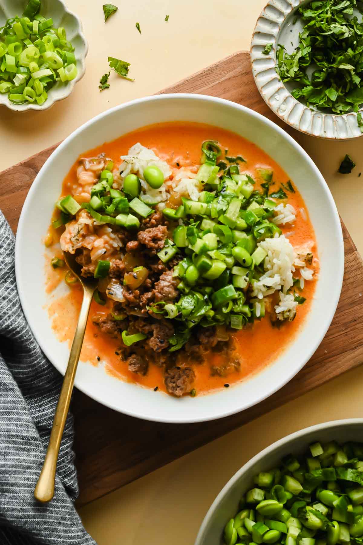 ground beef coconut rice bowl topped with edamame and fresh herbs in white serving dish with gold spoon.